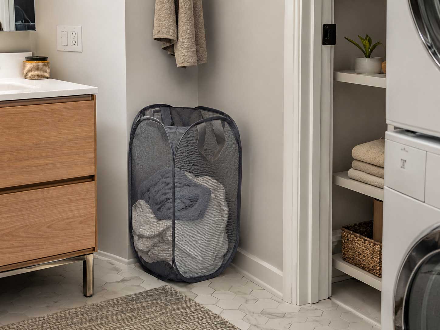 Collapsible mesh laundry hamper filled with clothes in a small apartment bathroom-laundry corner.